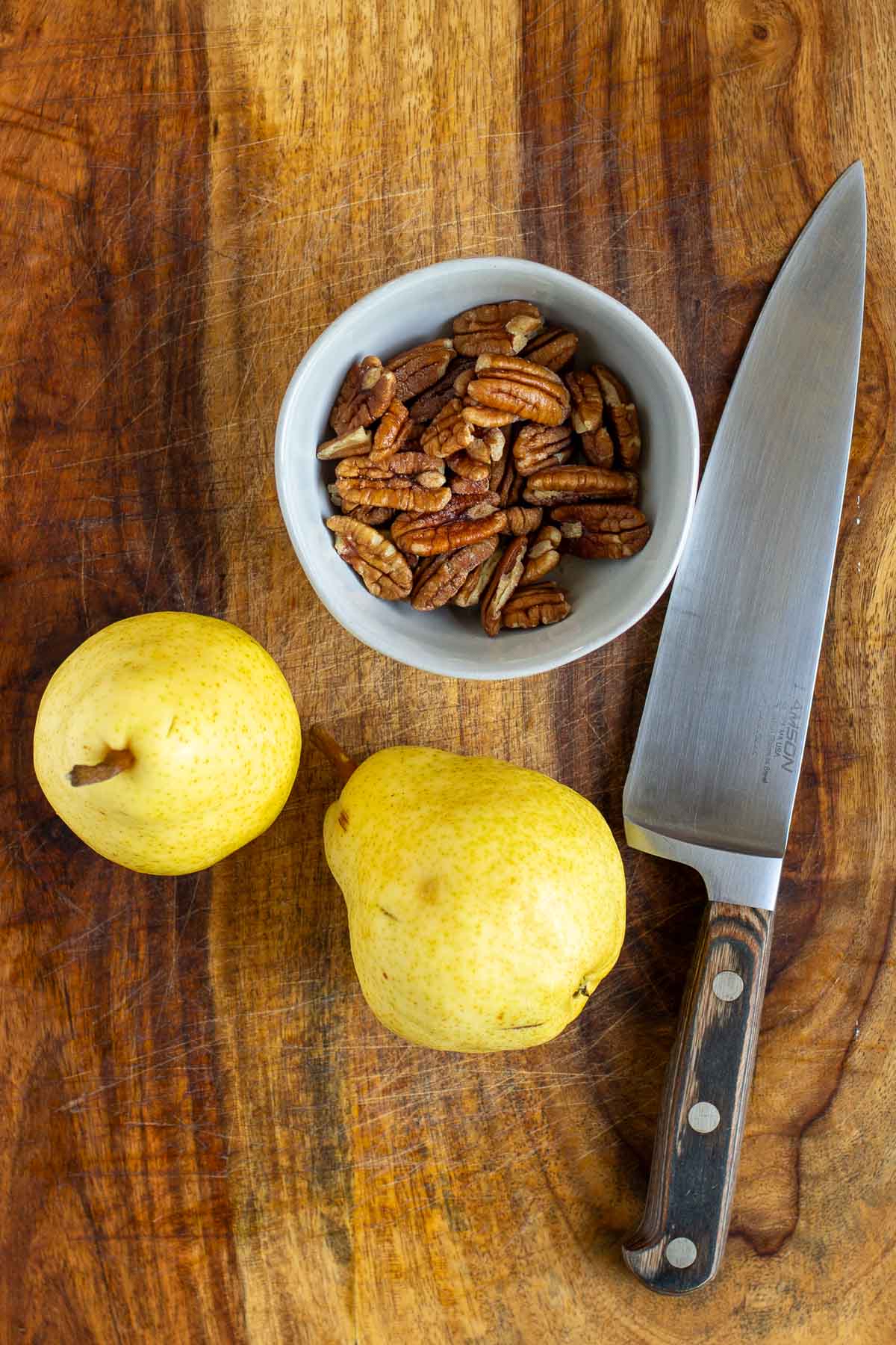 Top-down view of a well-worn wooden cutting board displaying two fresh yellow pears and a bowl filled with whole pecan nuts. A polished chef's knife with a wooden handle is placed to the right of the pears.