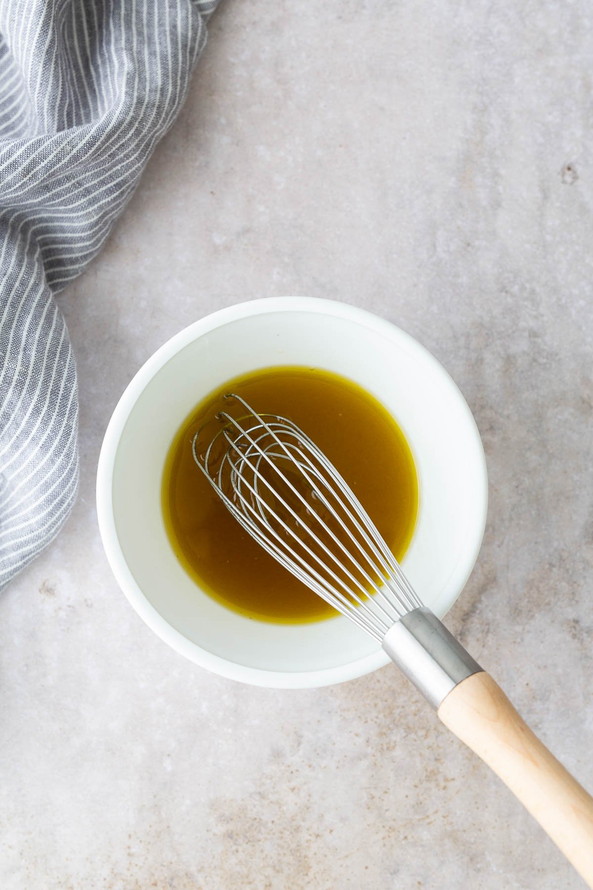 Overhead view of a white bowl with golden maple apple cider vinaigrette and a whisk on a light countertop, ready for a persimmon salad dressing.
