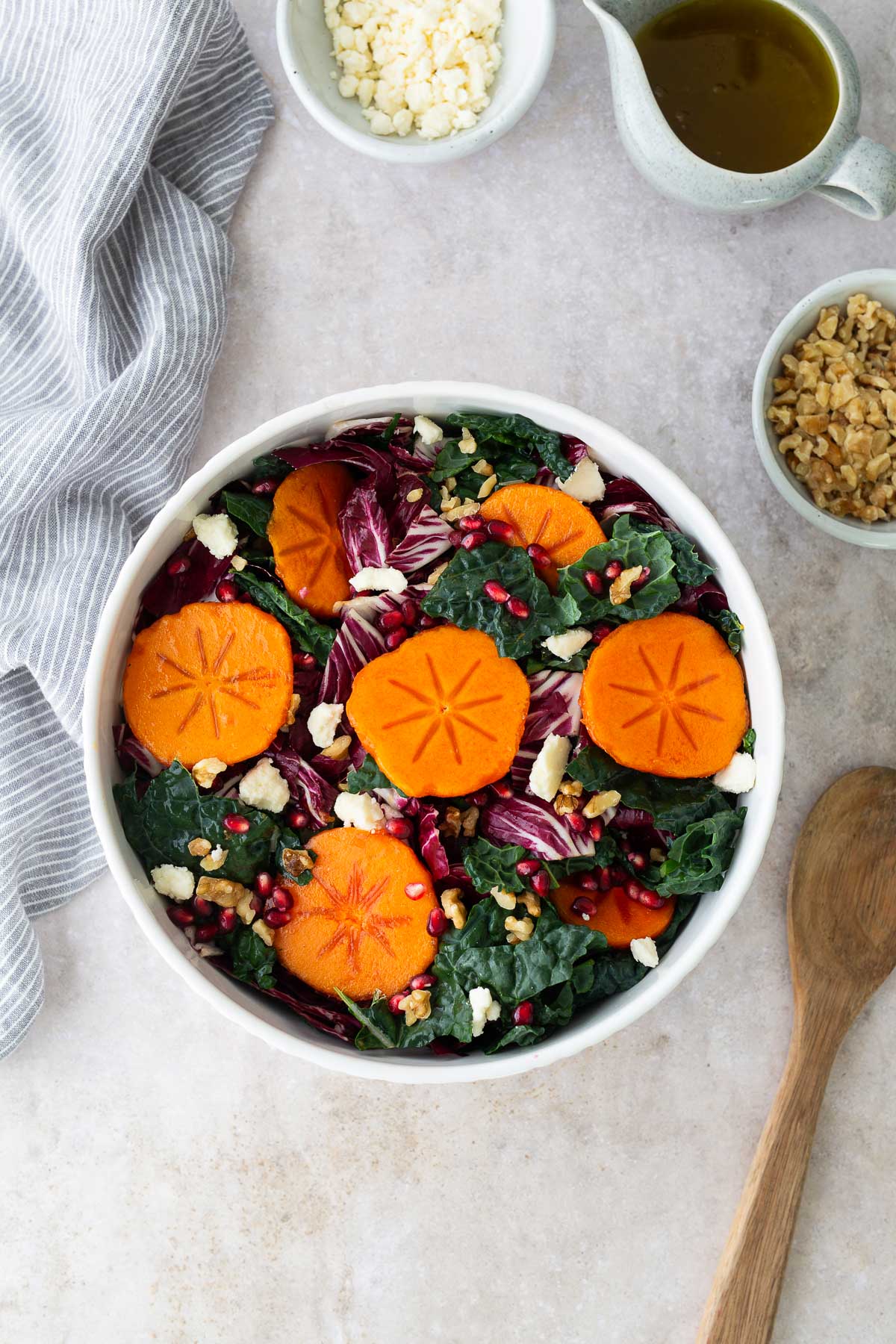 Fresh persimmon salad in a white bowl with sliced Fuyu persimmons, kale, radicchio, pomegranate seeds, feta, and walnuts surrounded by small bowls of ingredients