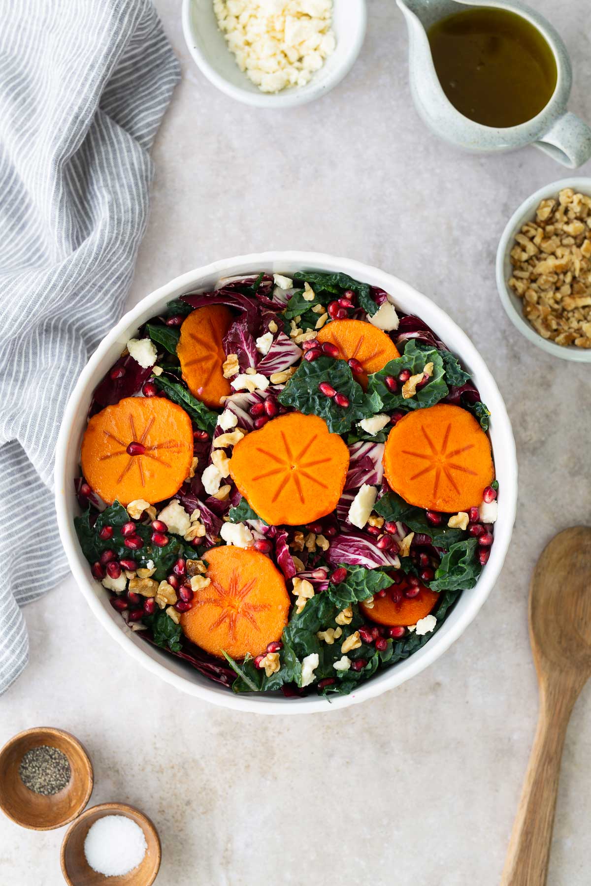 Colorful persimmon salad with kale, radicchio, pomegranates, walnuts, and feta cheese arranged in a serving bowl on a neutral countertop.