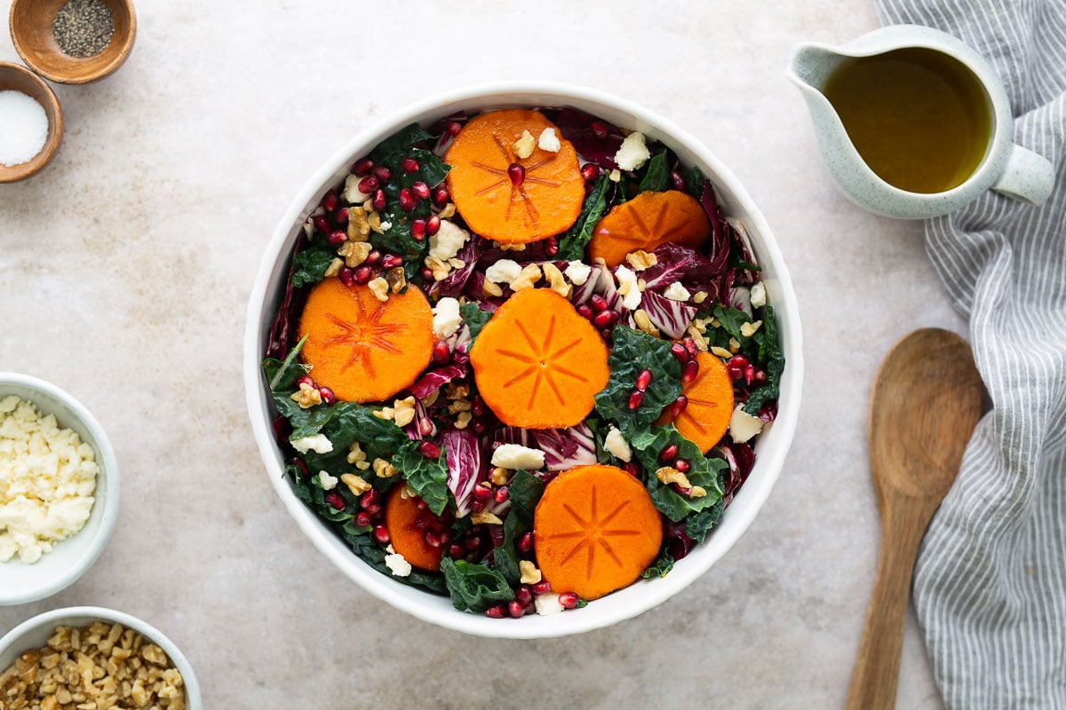Overhead shot of persimmon pomegranate salad in a white bowl, surrounded by salad dressing, feta, and walnuts for a vibrant fall salad presentation.