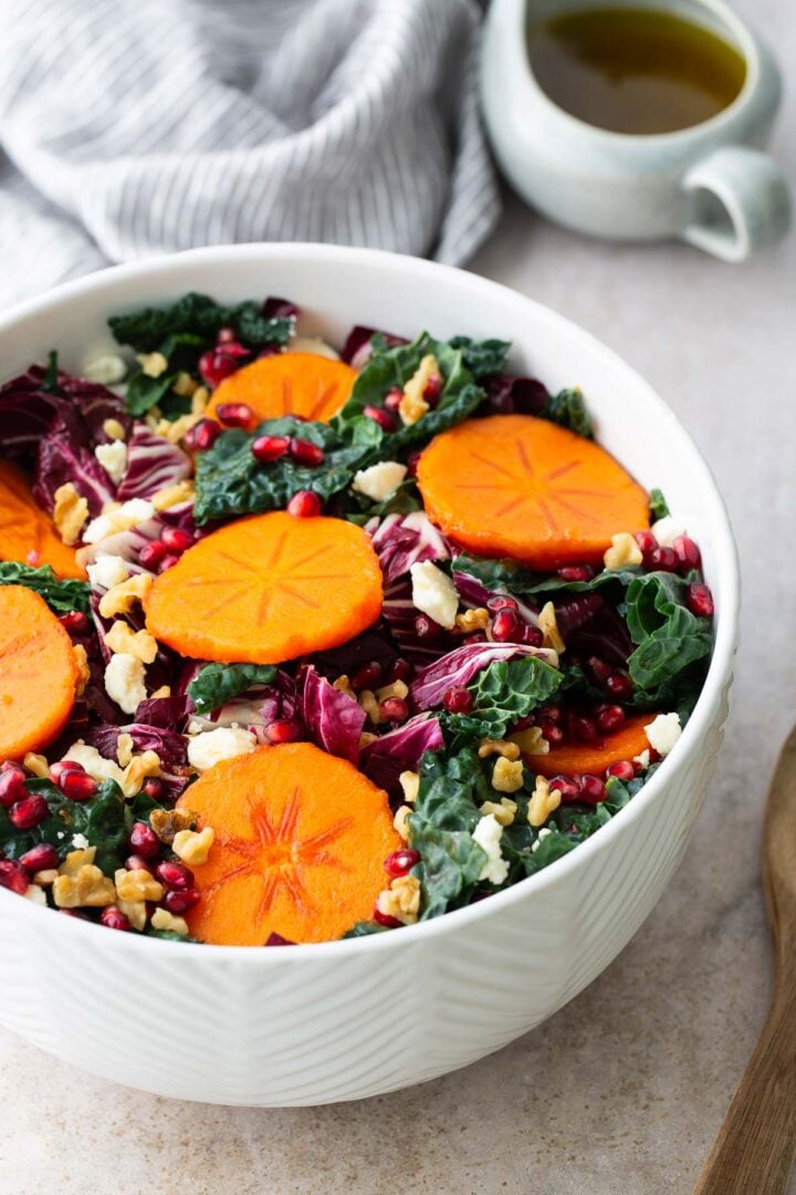 Side angle of a bright kale and persimmon salad with pomegranates, feta, and walnuts in a white textured bowl, perfect for a fall or holiday salad.