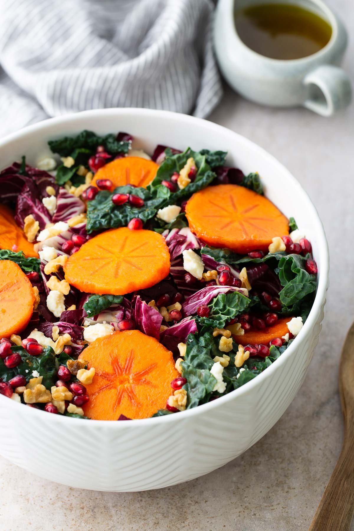 Side angle of a bright kale and persimmon salad with pomegranates, feta, and walnuts in a white textured bowl, perfect for a fall or holiday salad.