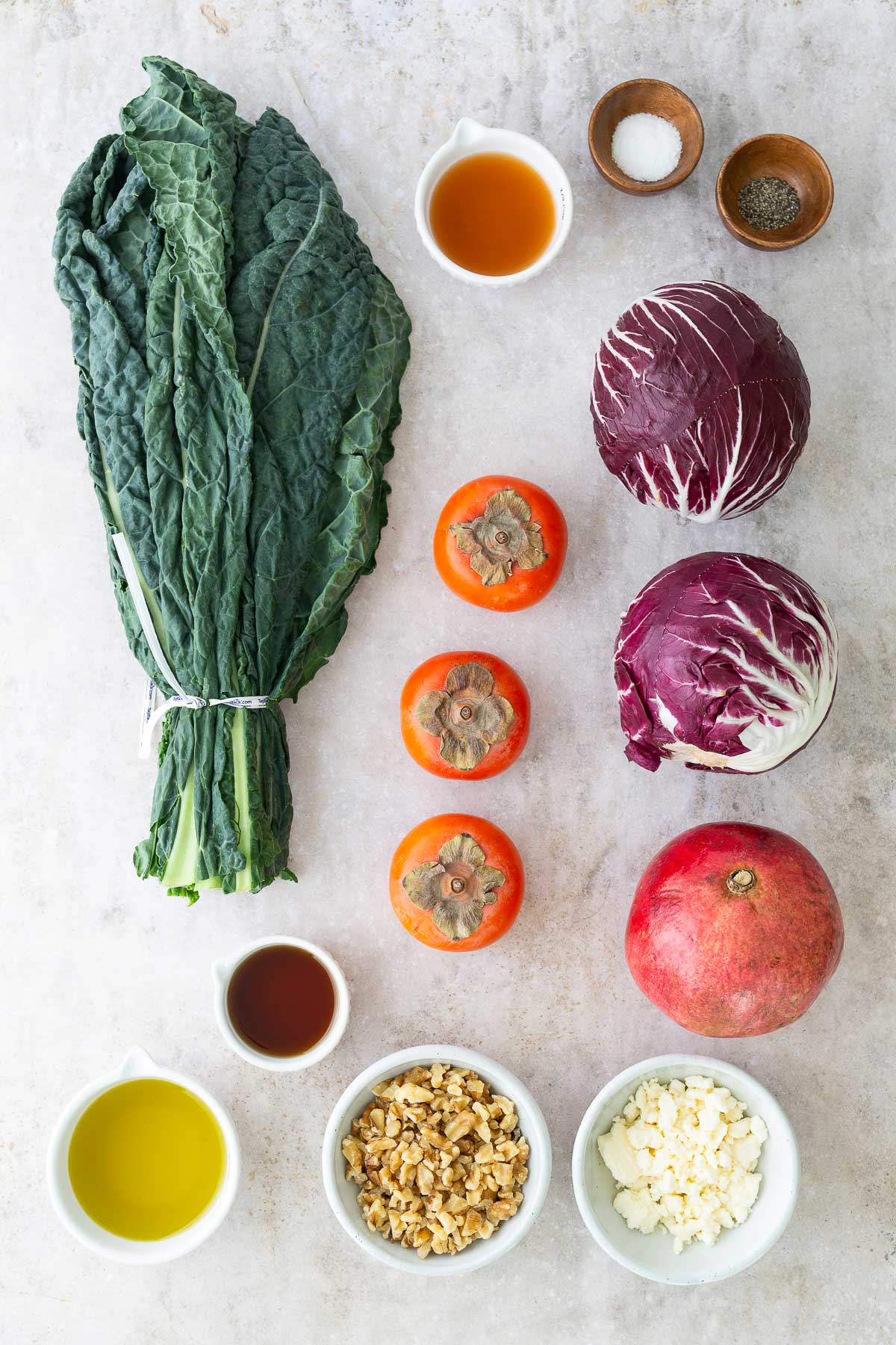 Overhead layout of fresh persimmon salad ingredients including kale, radicchio, Fuyu persimmons, pomegranate, feta, walnuts, olive oil, vinegar, and maple syrup on a light background.