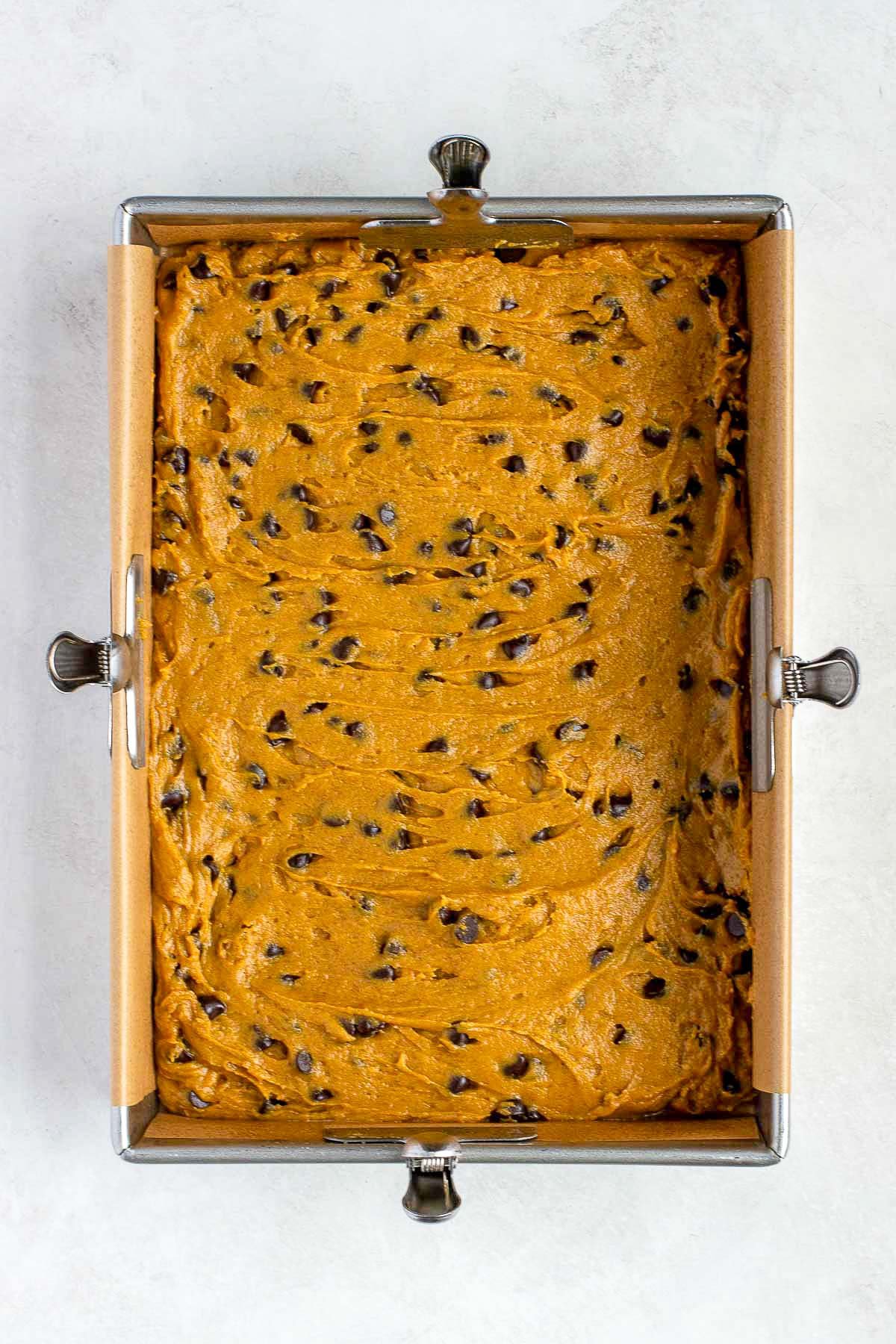 Pumpkin chocolate chip cookie dough spread evenly in a parchment-lined baking pan before baking.