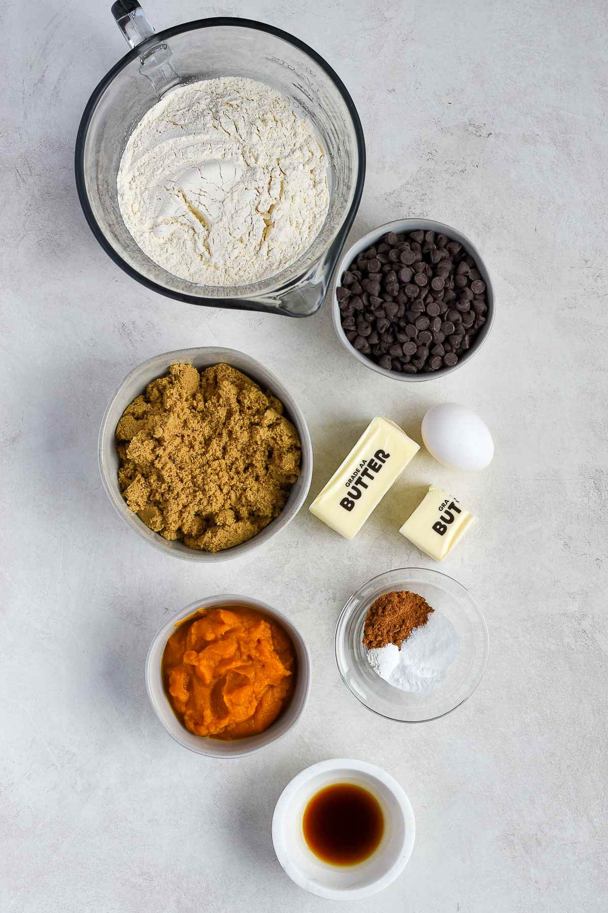 Overhead shot of ingredients for pumpkin chocolate chip cookie bars including flour, butter, brown sugar, pumpkin purée, chocolate chips, egg, and spices arranged on a light gray surface.