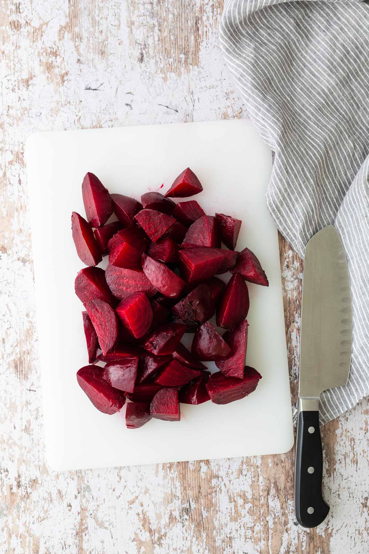 Chopped roasted beets on a cutting board with a chef’s knife and striped towel on rustic wood surface.