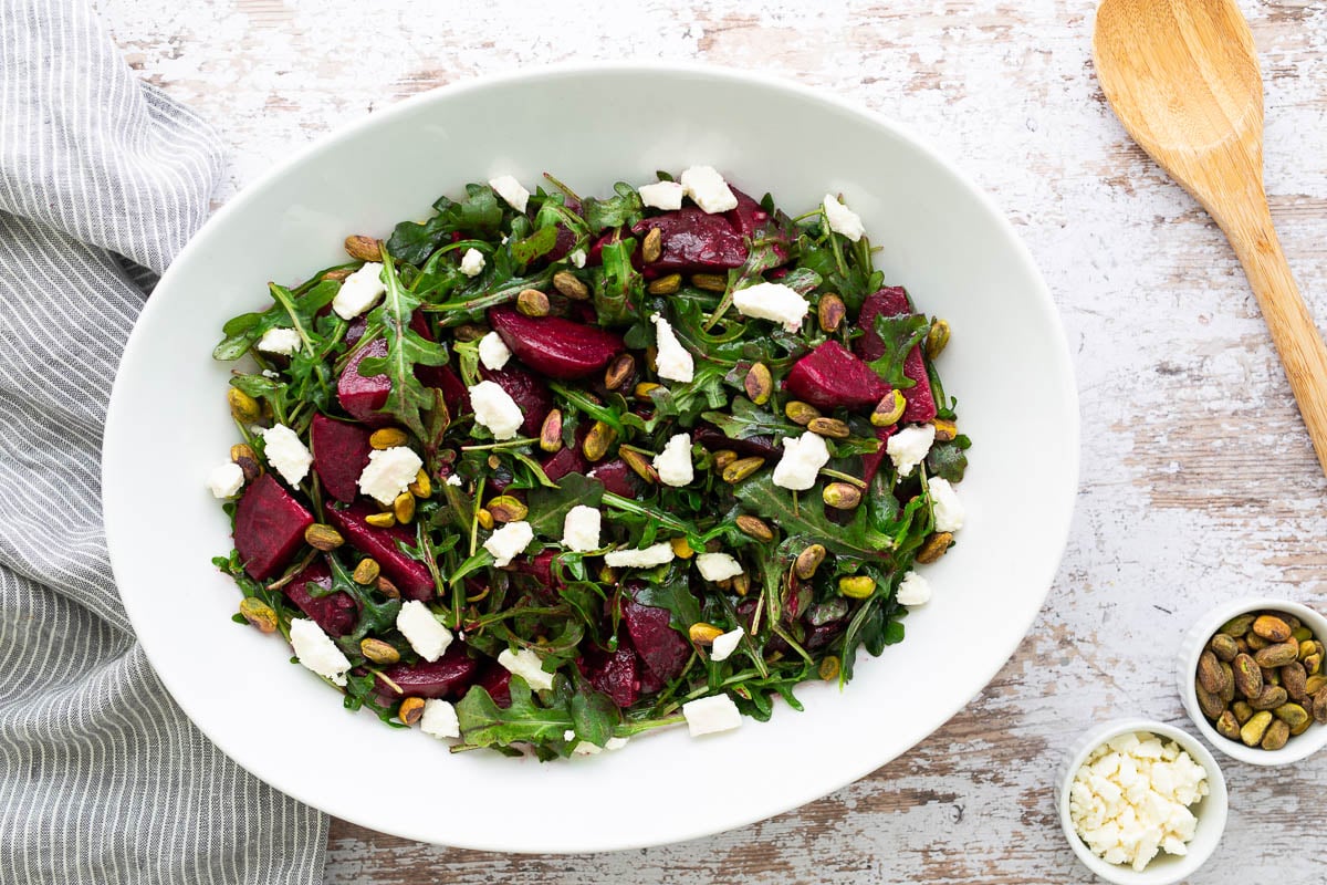 Overhead view of finished roasted beet salad with feta and pistachios in white bowl with small bowls of cheese and nuts on the side.