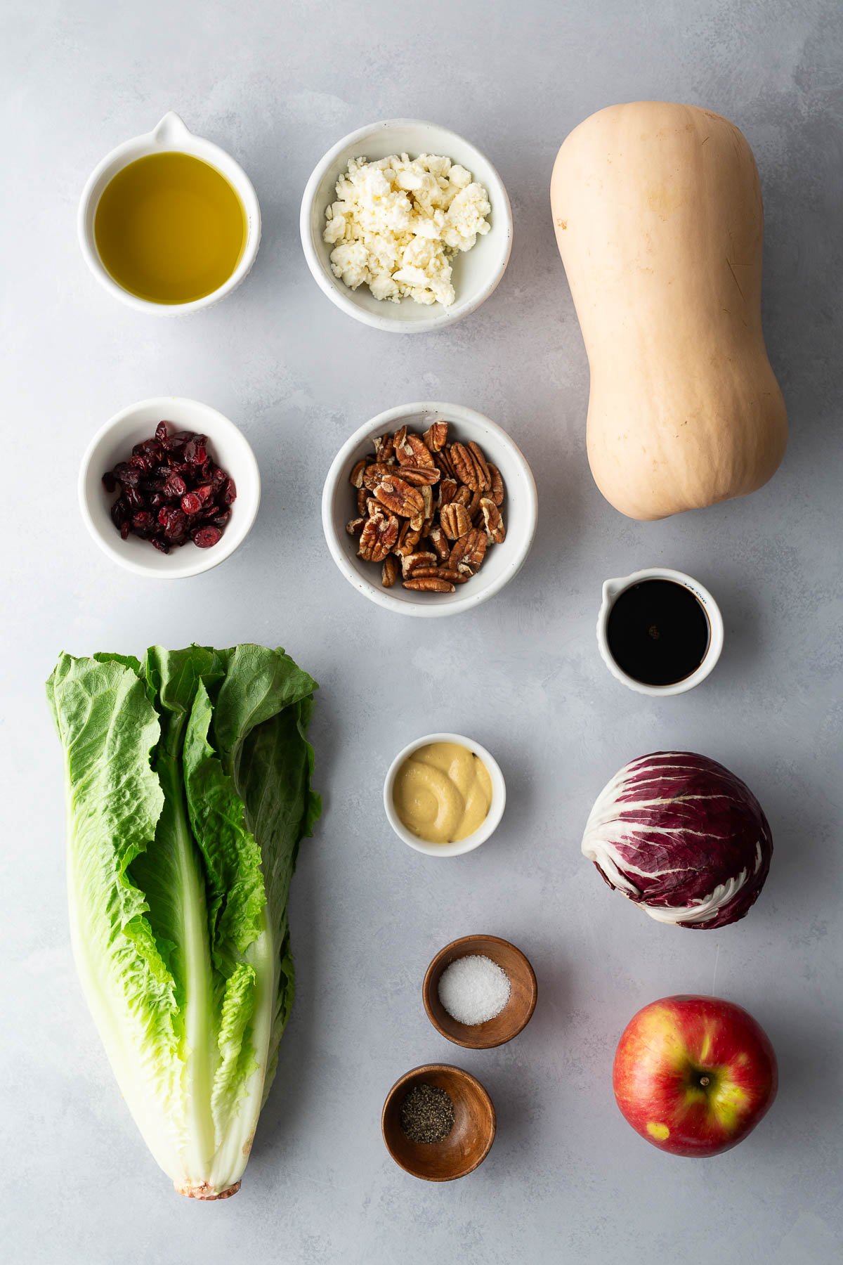 Ingredients for roasted butternut squash salad with feta, pecans, apple, and balsamic dressing on a light gray surface.