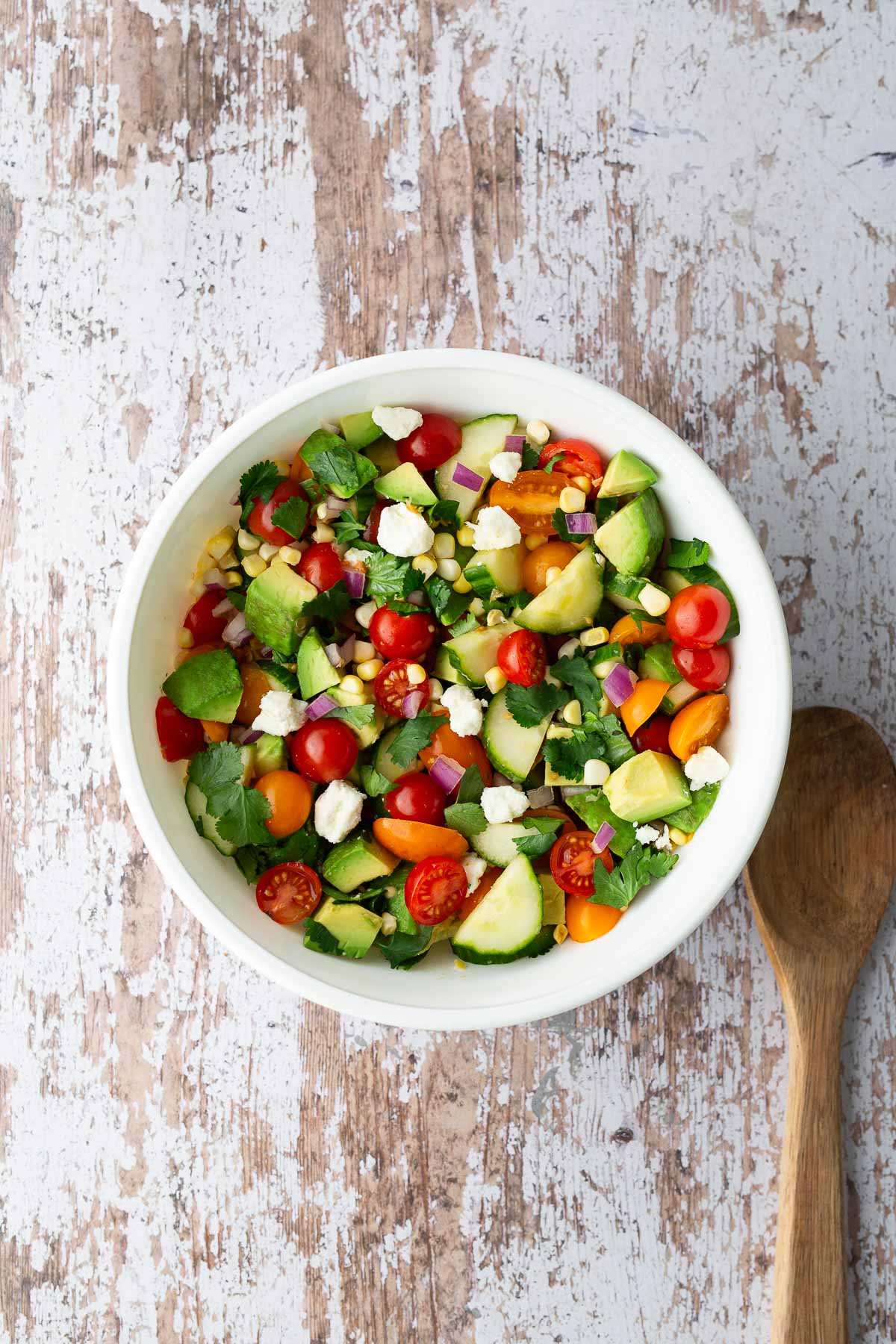 Tomato, corn, cucumber, avocado salad in a white bowl with feta, red onion, and cilantro.