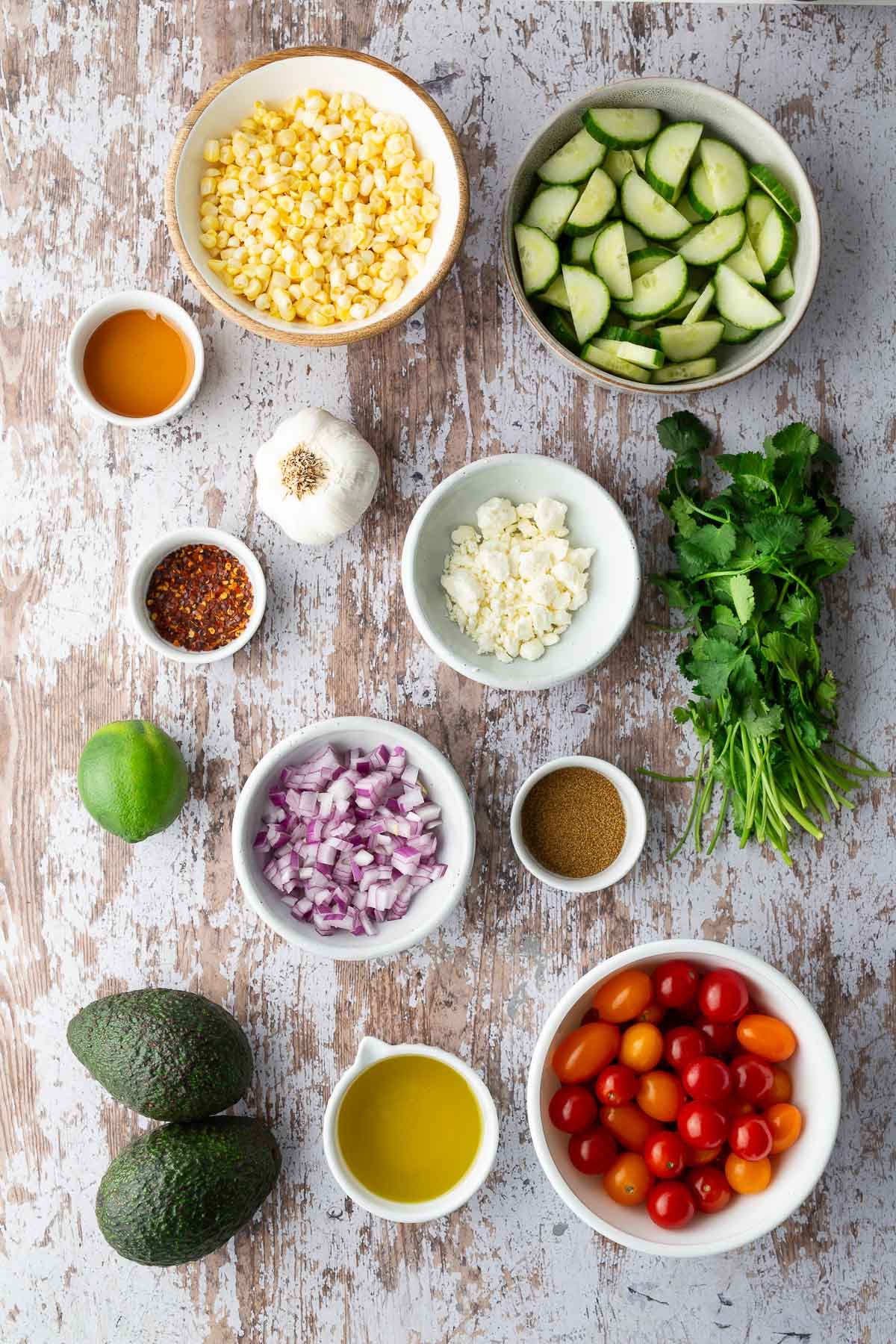 Flat lay of fresh salad ingredients: corn, cucumber, avocado, cherry tomatoes, red onion, cilantro, feta, lime, and seasonings—for tomato corn cucumber avocado salad recipe.